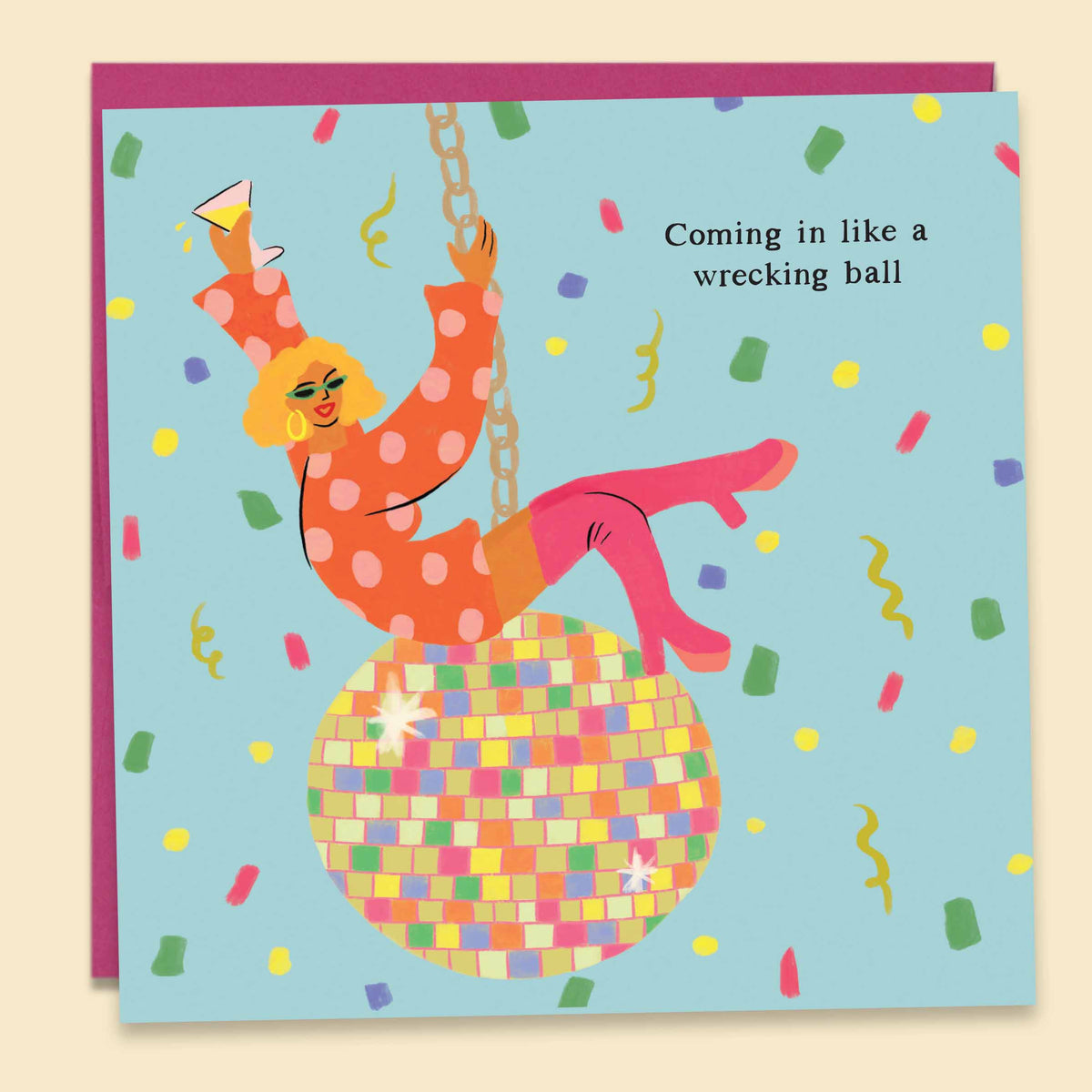 Illustrated by Constanza Goeppinger a greeting card featuring a woman in a party outfit riding a colourful disco ball holding a glass of wine, surrounded by bright confetti with the text 'Coming In Like A Wrecking Ball '. Australian-made greeting card by Papernest