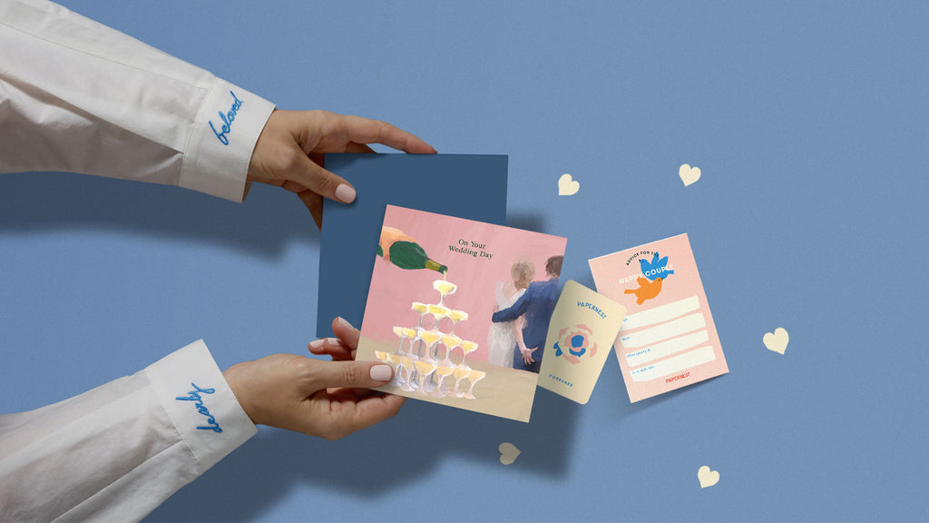 Hands holding funny Australian wedding card with champagne tower illustration, plantable heart confetti, and Wishes for the Happy Couple insert. Embroidered cuffs read 'dearly beloved