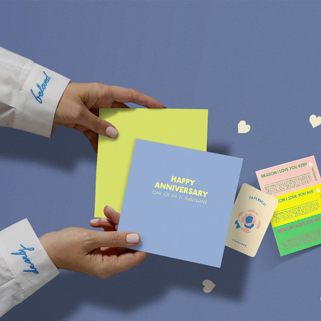 Hands holding funny anniversary card that says Happy Anniversary One of us is punching with yellow envelope, plantable seed paper confetti hearts, and novelty fortune insert on blue background, white shirt cuffs embroidered with Dearly Beloved