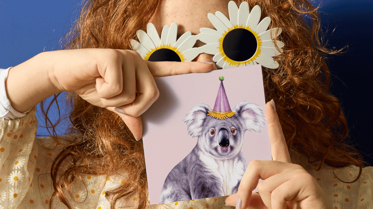 A person with curly red hair holds a square card featuring a koala wearing a party hat against a pink background. The person is wearing white daisy-shaped sunglasses and a cream floral top, posed against a dark blue background.