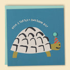 Greeting card with an illustration of a turtle wearing a party hat and balloon, with the text 'Have a Turtley AWESOME DAY!' The card is also decorated with a pom-pom detail  — Australian-made greeting card by Papernest