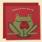 A greeting card with a frog illustration, with a real red heart balloon tongue inserted in its mouth, and the text 'TOADALLY IN LOVE WITH YOU' on the top. Australian-made greeting card by Papernest.